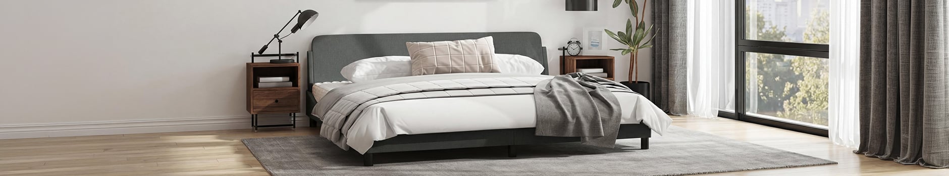 Vue large d’une chambre moderne avec un cadre de lit gris foncé, du linge de lit blanc et un coussin beige à motif quadrillé. La pièce comprend deux tables de chevet en bois avec lampes, une grande plante en pot et de grandes fenêtres avec des rideaux gris et blancs donnant sur des arbres verts.