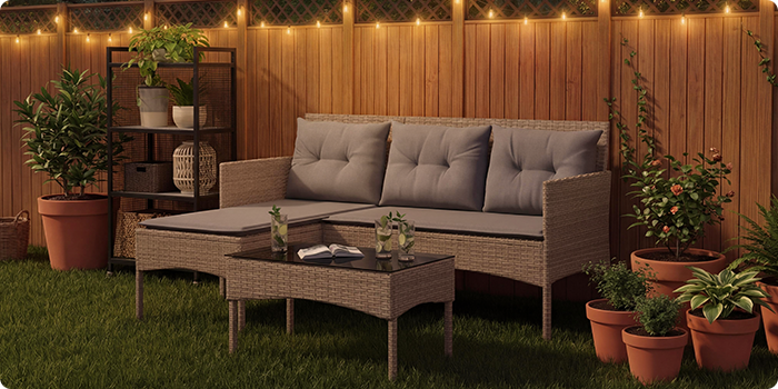 Salon de jardin lounge en rotin avec des coussins gris, comprenant un canapé trois places, un repose-pieds et une table basse sur une pelouse avec des guirlandes et des plantes