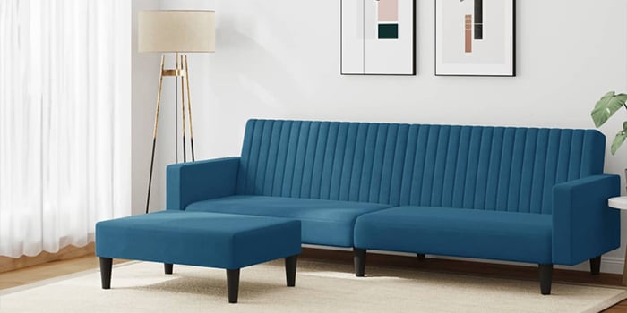 Canapé d’angle moderne en velours bleu avec capitonnage vertical et pouf assorti. Le canapé possède des pieds en bois noir et se trouve dans un salon lumineux, avec des tableaux encadrés au mur et un lampadaire dans le coin.